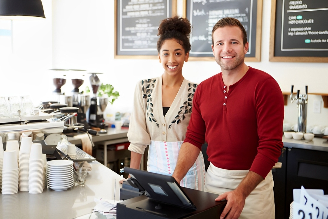 Two friendly baristas smiling behind counter in a bright coffee shop.