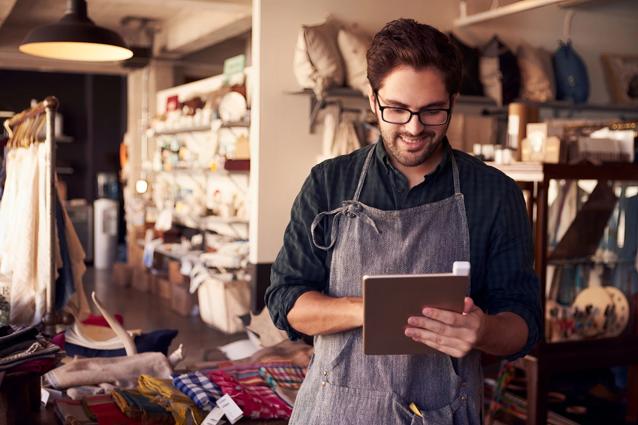 Shop owner in apron using tablet while standing inside a boutique.